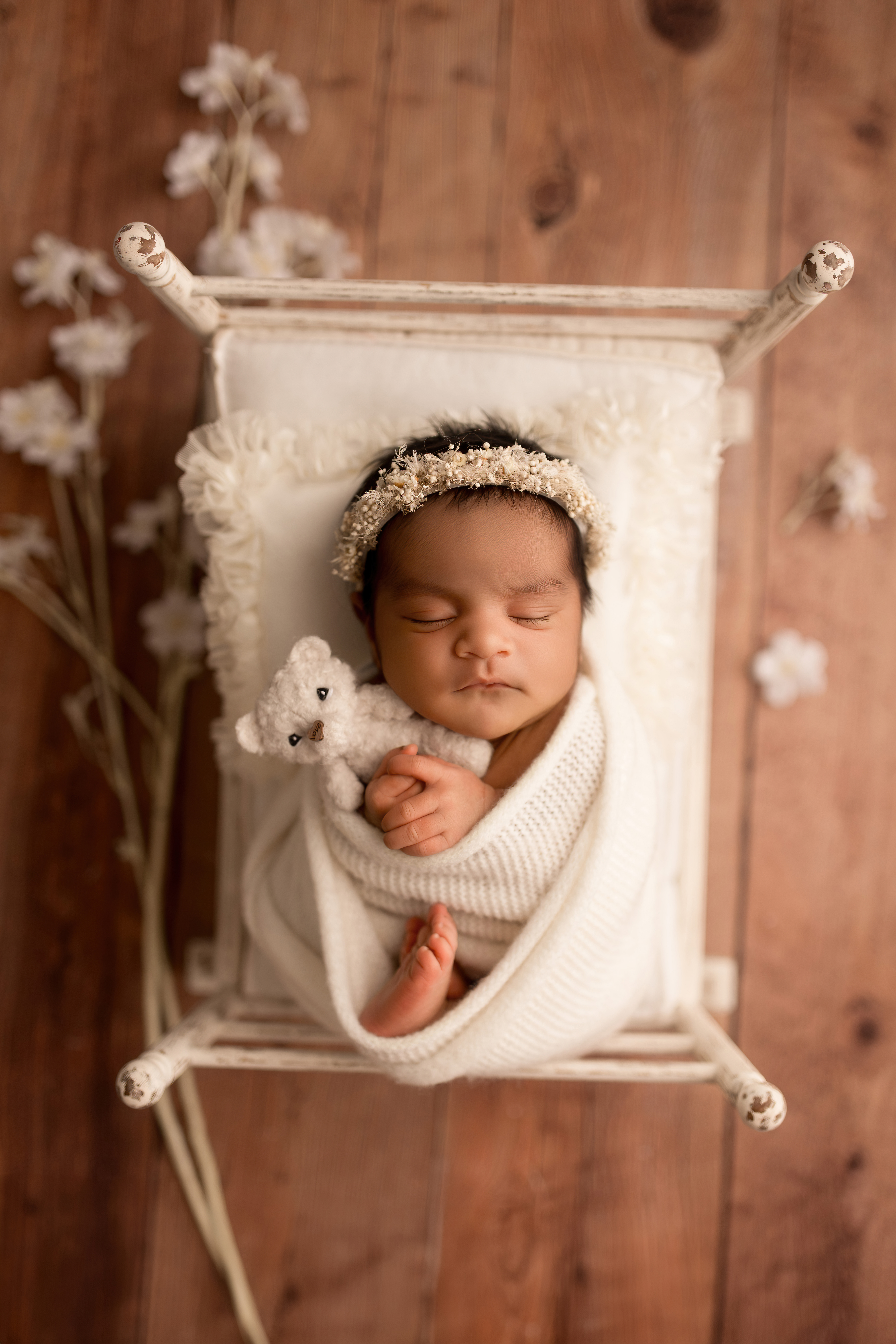 A sleeping newborn wrapped in a cozy white blanket, cradling a small stuffed animal, surrounded by delicate flowers on a wooden background.